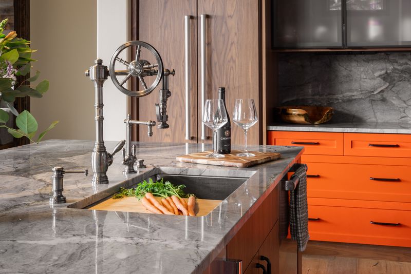 Kitchen island with wheel-handle bridge faucet, quartzite counter, undermount sink, and orange cabinets beyond