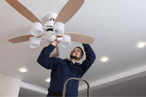 A technician is adjusting a ceiling fan while standing on a ladder.
