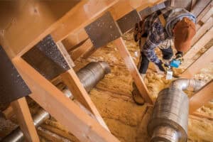 A worker in a cap is inspecting ductwork inside an attic, surrounded by insulation and wooden beams.