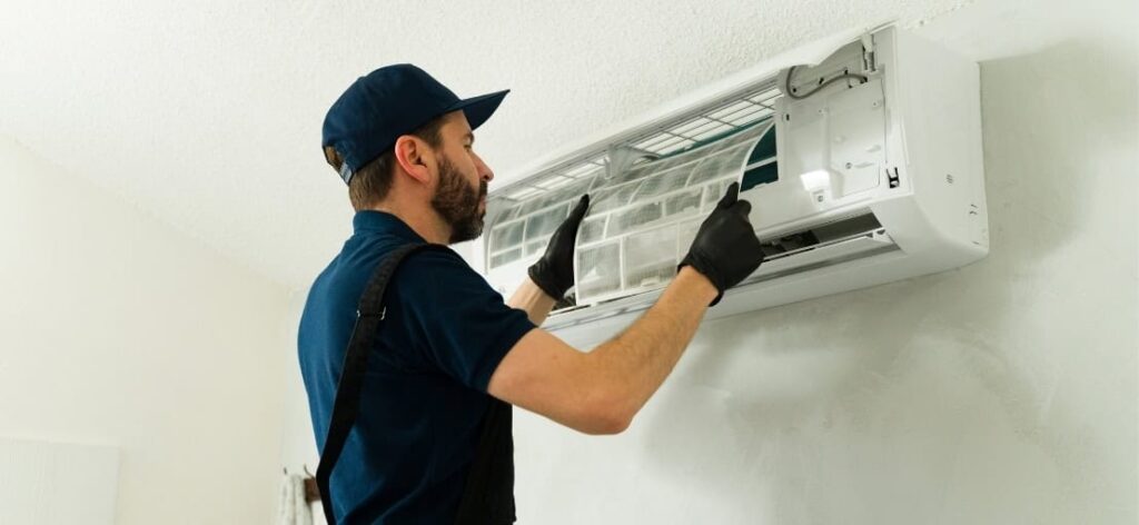 A technician in a blue uniform is removing a filter from an air conditioning unit mounted on a wall.
