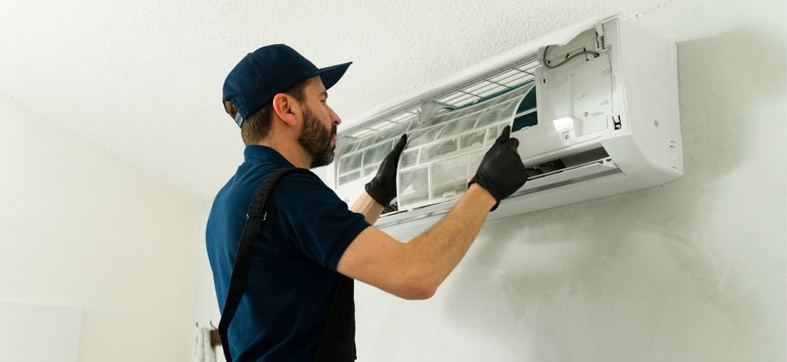 A technician in a blue uniform is removing a filter from an air conditioning unit mounted on a wall.