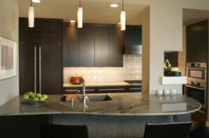 G-shaped kitchen with dark, wooden cabinets, subway tile walls, and a grey quartz countertop bar with three black bar chairs.