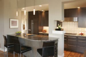 G-shaped kitchen with dark, wooden cabinets, subway tile walls, and a grey quartz countertop bar with three black bar chairs.