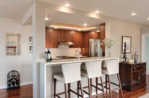 Small condo kitchen alcove with wooden cabinets, and a white bar countertop and three white bar stools.