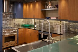 U-shaped contemporary kitchen with grey subway tile walls, black quartz countertop, chrome appliances, and light-wooden cabinets.