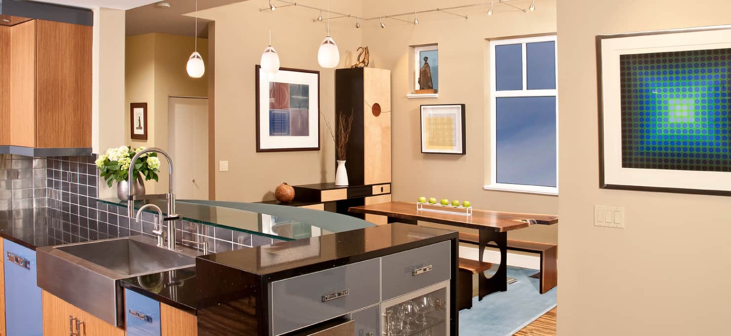 Contemporary kitchen overlooking a dining area with tan walls, table with wooden benches, and contemporary art on the walls.