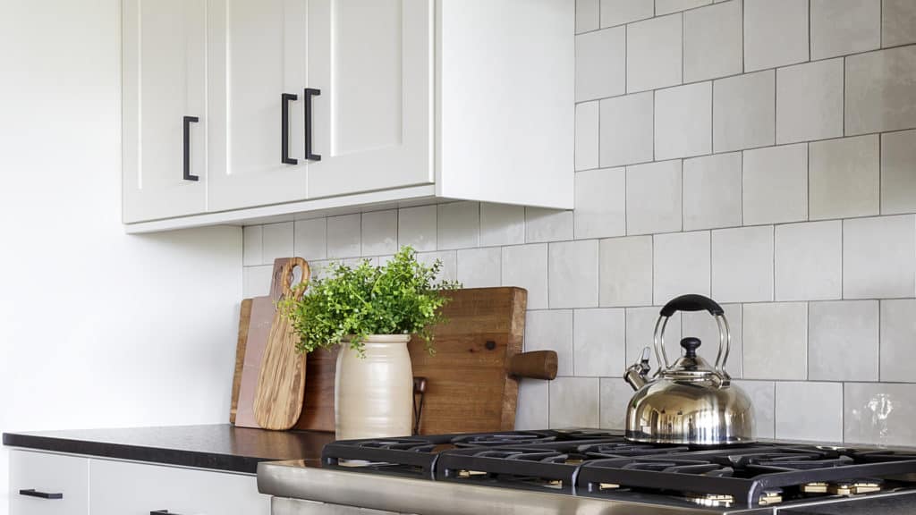 Cloe handmade light grey ceramic tiles in a modern kitchen with white cabinets, a black countertop, and stainless steel gas stove. 