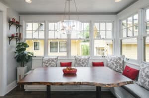 An eclectic dining room has a white banquette beneath large windows, wood table, antique chairs, and contemporary metal light fixture.