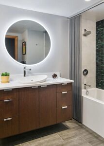 Mahogany vanity with a white countertop beneath a round, backlit mirror next to an alcove bathtub.