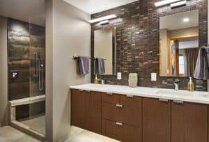 Mahogany vanity with white countertops beneath two mirrors with overhead lights on a dark tiled wall.