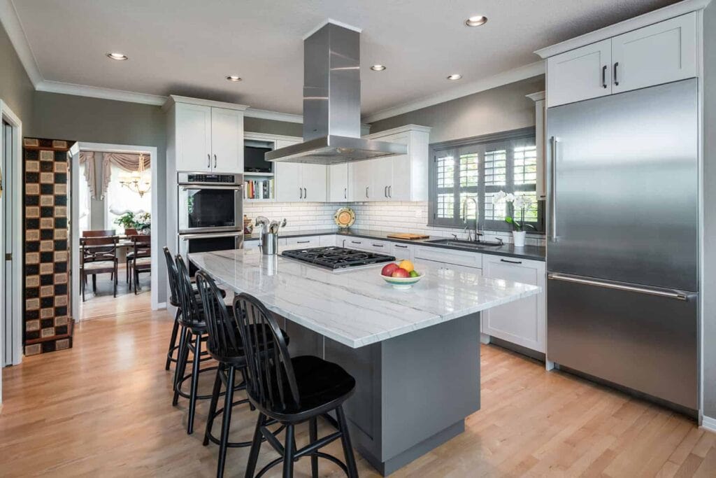 Modern kitchen with a quartzite island with a gas range, stainless steel air vent, and four black bar chairs.