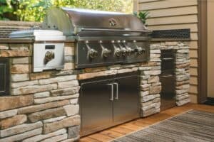 Stylish outdoor grill made of stainless steel and stone, featuring multiple burners and storage underneath a wooden structure.