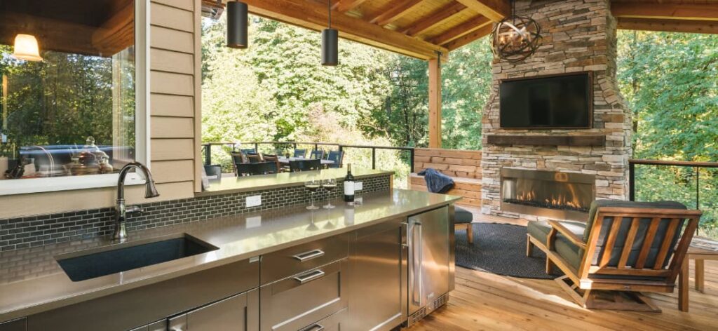 Open outdoor kitchen with stainless steel appliances, a cozy seating area, and a stone fireplace surrounded by greenery.