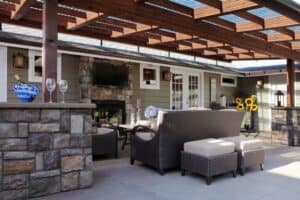 Cozy outdoor seating area with stone accents, comfortable furniture, a stone fireplace, and a TV under a wooden pergola.
