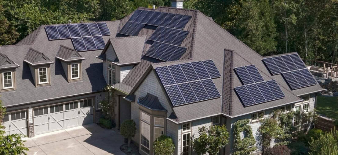 A large house with multiple solar panels installed on its roof, surrounded by trees and a landscaped yard.