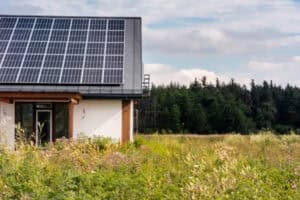 A side view of a modern house with solar panels on the roof, set in a field with tall grass and a forest in the background.