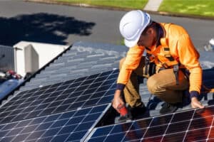 A technician in a hard hat is working on solar panels atop a roof, using a drill to adjust equipment.
