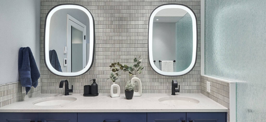 Dual modern bathroom sinks with oval mirrors, grey tiled wall, potted plant, black fixtures, navy towels, and frosted glass panel.