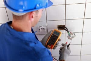 Electrician inspecting wiring behind tiled wall, wearing blue uniform and safety helmet, using a multimeter for electrical testing.