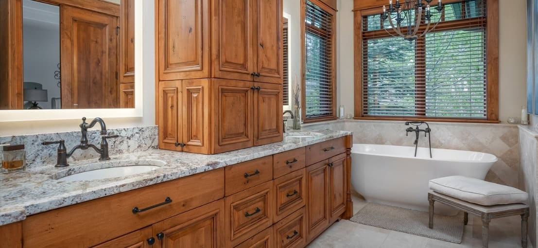 A luxurious bathroom with wood vanity, granite countertops, dual sinks, a freestanding tub by a window, and a cushioned stool.