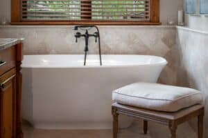 Elegant bathroom with a freestanding tub by a window, wooden vanity, granite counters, and a cushioned stool.
