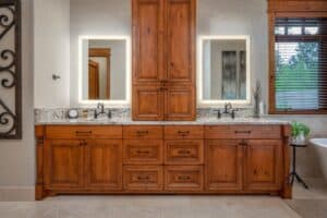 Double-sink vanity with granite counters, warm wood cabinets, framed mirrors, and a window backdrop.