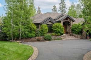 A suburban home with a shingled roof, stone porch, and lush landscaping, surrounded by trees.