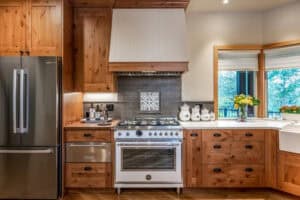 Kitchen featuring natural wood cabinetry, a stainless steel side-by-side refrigerator, gas stove with a white range hood, stone tile backsplash, and a light-colored countertop.