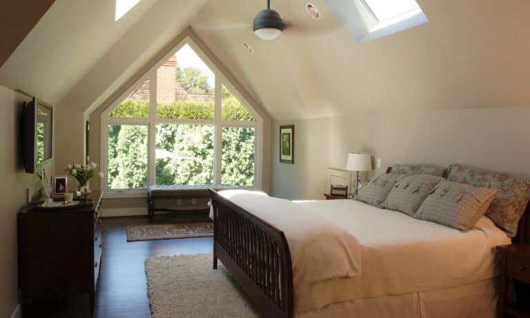 Bright attic bedroom with a dark wood sleigh bed, shag rug, and large A-frame window overlooking greenery..)
