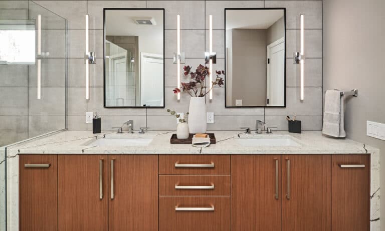 Modern primary bathroom with double vanity, wood cabinets, light gray tile, and two mirrors.)