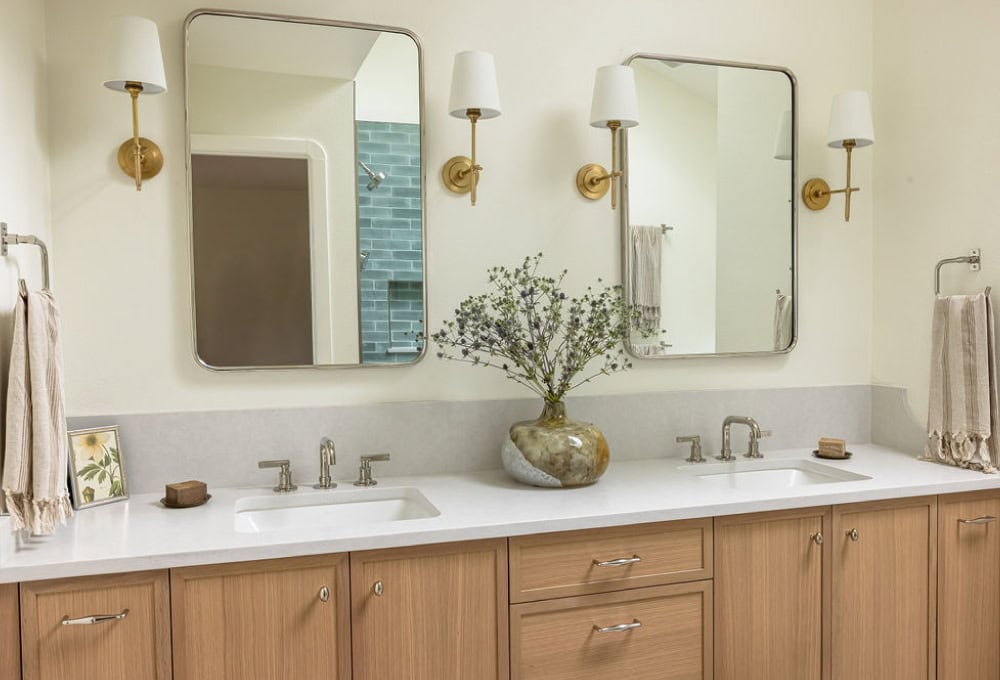 A traditional bathroom vanity with light wood cabinets, a marble countertop, a framed mirror, two wall sconces, and a small decorative plant on the counter.