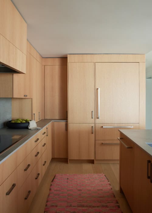 A minimalist kitchen with light wooden cabinetry, sleek countertops, and a refrigerator integrated into the cabinetry, complemented by a pink rug.