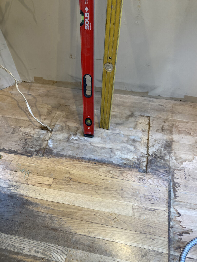 A hardwood floor with visible moisture damage and tools leaning on the wall