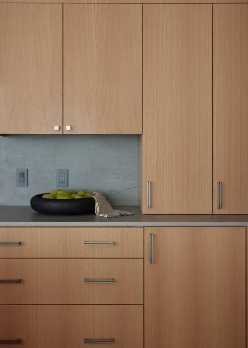 A close-up of a kitchen countertop displaying a dark bowl filled with yellow-green fruits, accented by light wooden cabinetry and a woven basket.