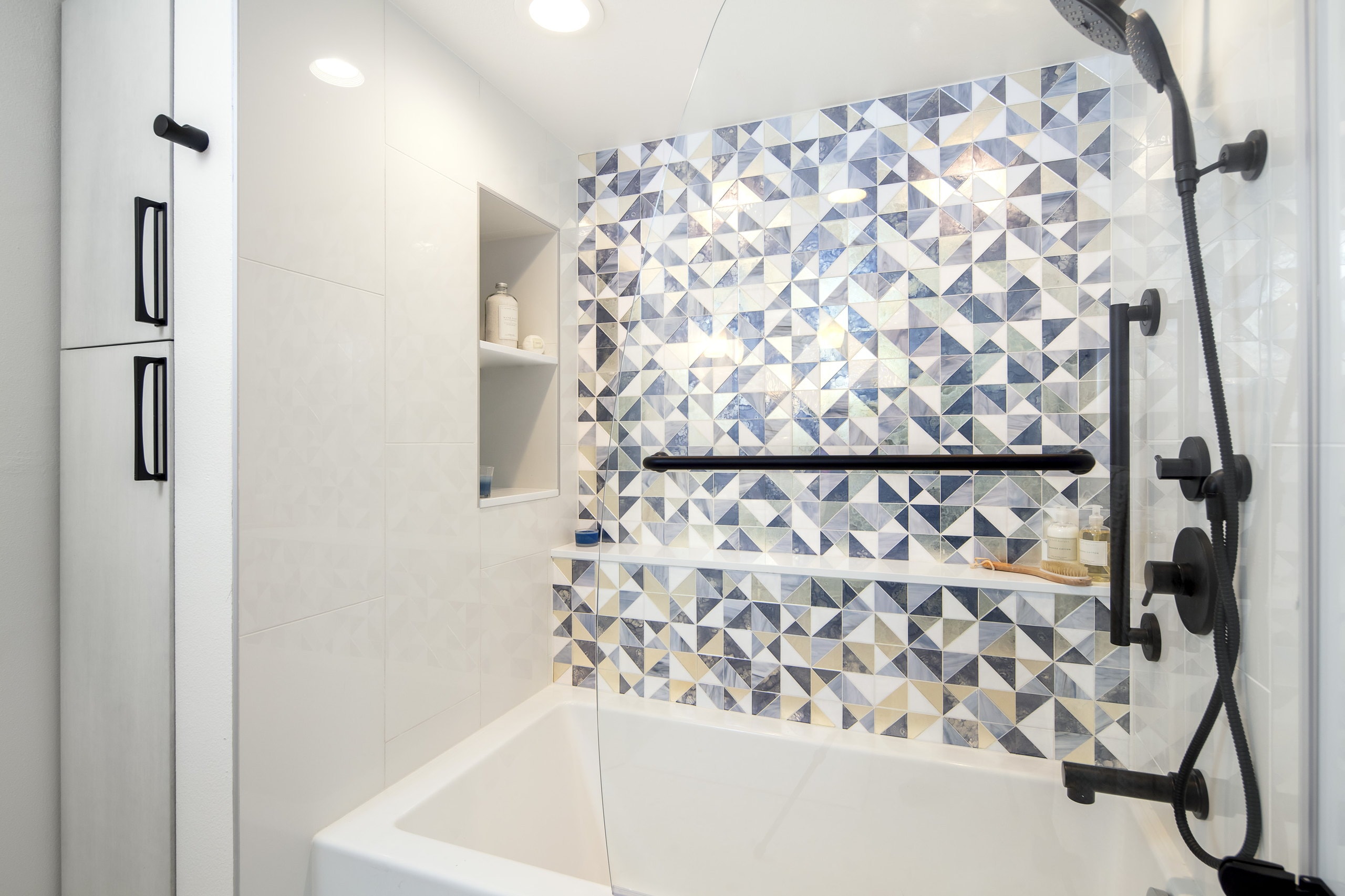 Alcove tub and shower faced in a blue, green, and cream geometric glass mosaic, with matte black fittings and a grab bar.