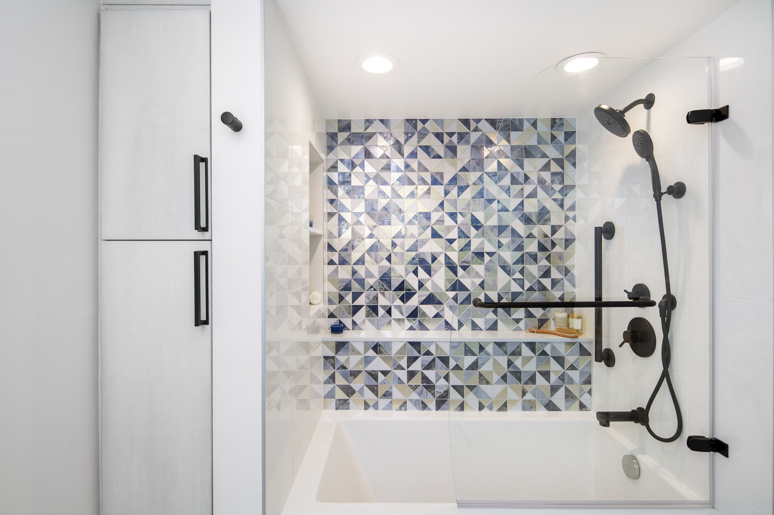 Tub and shower with patterned glass mosaic tile, a full-height linen tower at left, and a fixed glass splash panel.