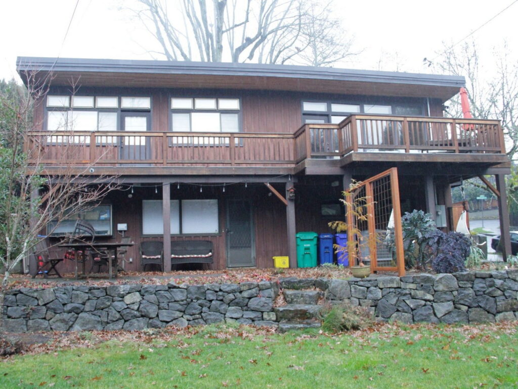 The house before remodeling, with dark brown stained siding and a turned-baluster wood deck rail, photographed on an overcast day.