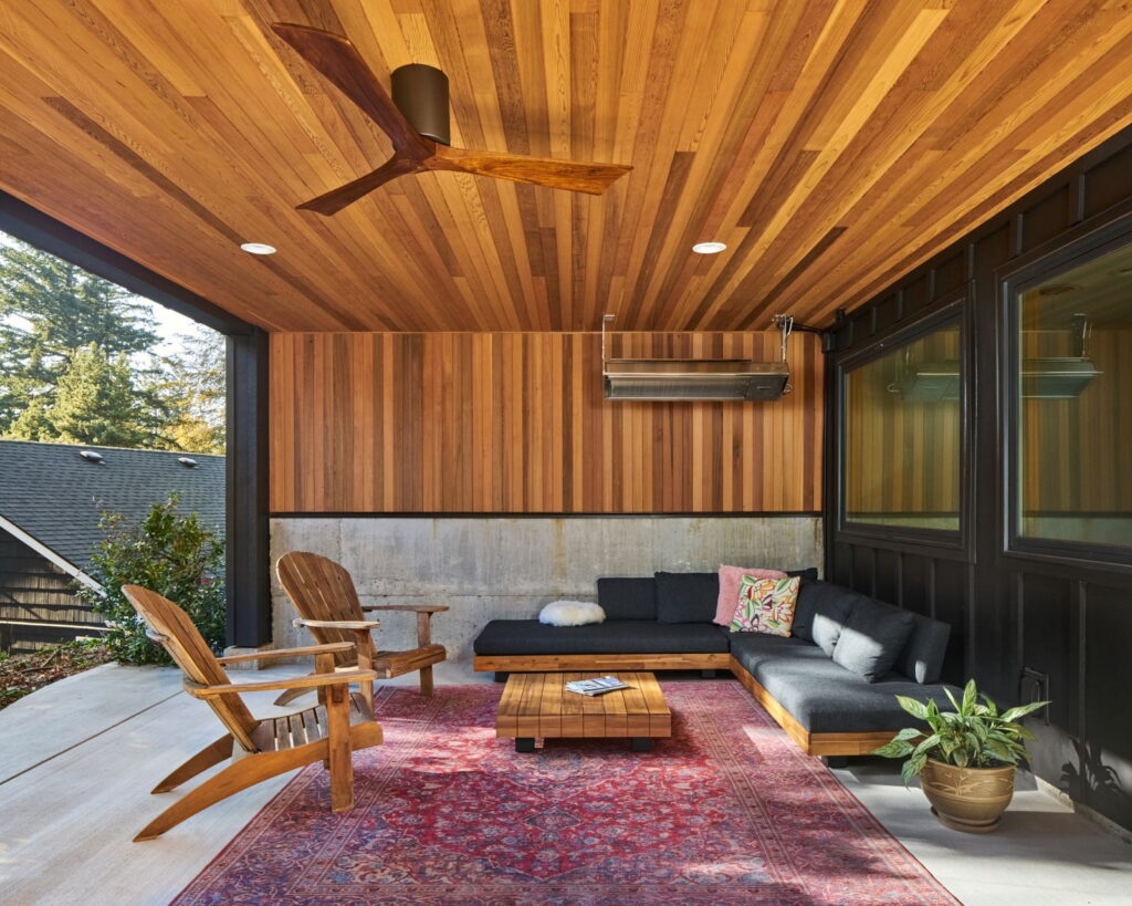 Covered outdoor room with a cedar plank ceiling, ceiling fan, overhead radiant heater, and board-formed concrete wall.