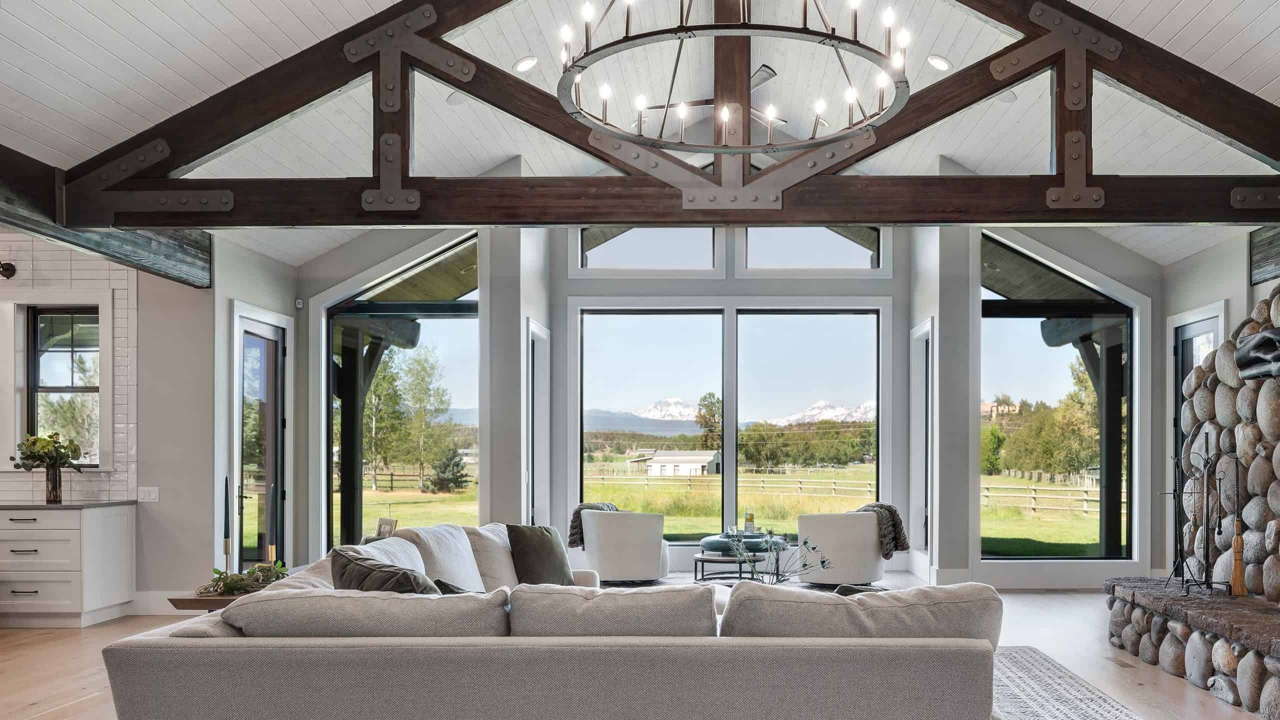A Central Oregon farmhouse living room with exposed trusses, a chandelier, a sectional sofa, and large windows with a view of the countryside.