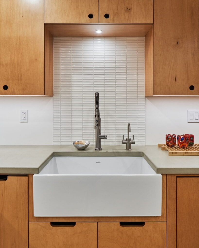 White fireclay apron sink below a niche of stacked white tile, set into concrete countertops.