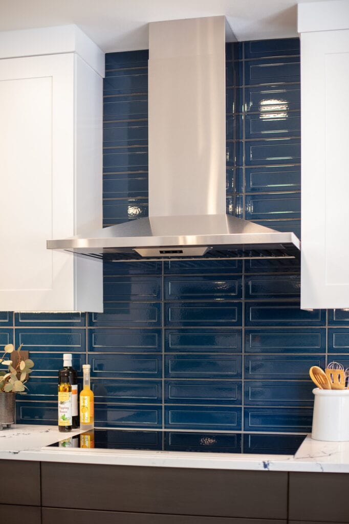 Stainless chimney hood centered on the glossy navy picket tile behind the cooktop.