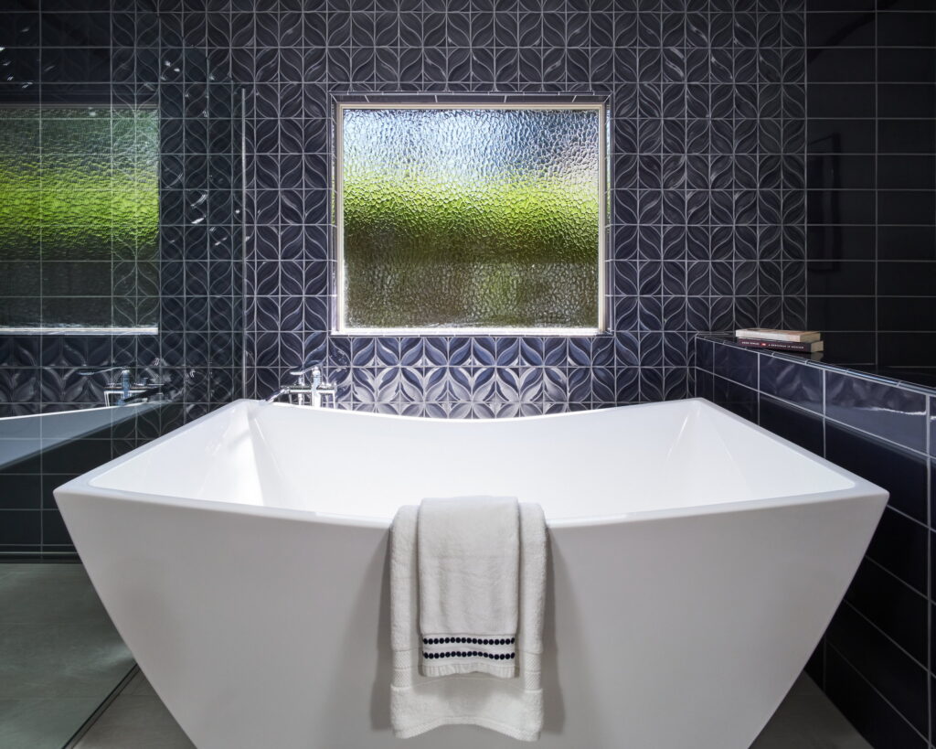 Freestanding angular tub centered beneath a textured glass window, framed by navy relief tile.