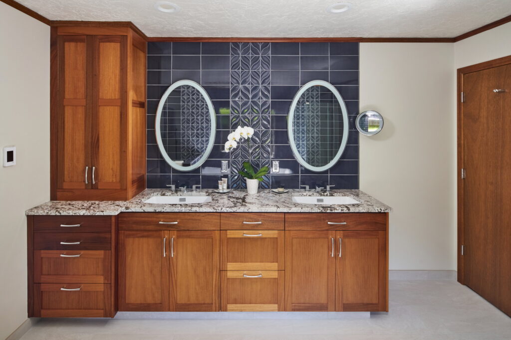 Mahogany double vanity with speckled granite counters and two backlit oval mirrors on the navy tile wall.