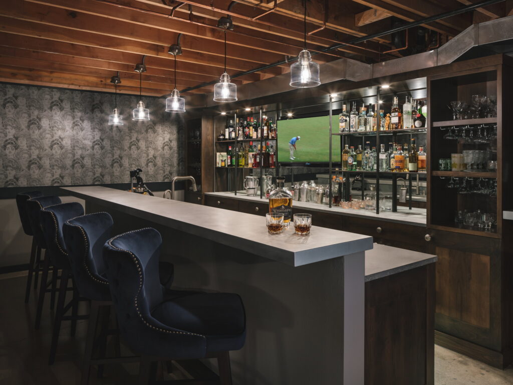 The finished bar with a honed concrete counter, navy velvet stools, and patterned wallpaper along the back wall.