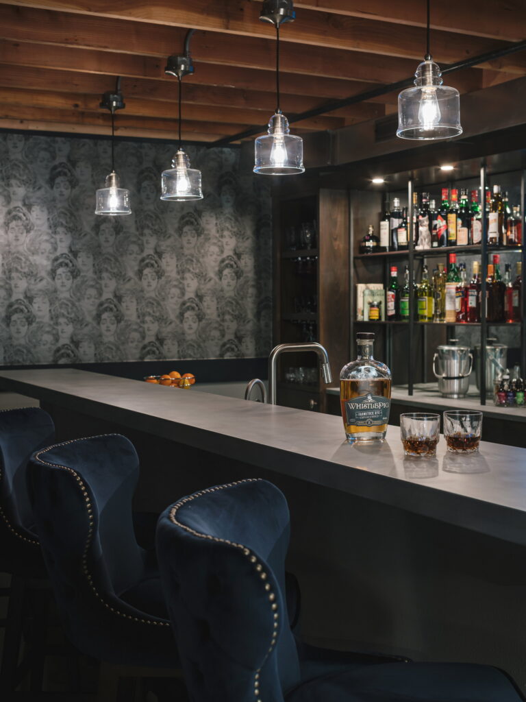 Bar counter set with a whiskey bottle and two glasses, patterned wallpaper behind.