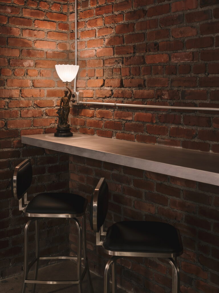 Stainless rail-height counter and two steel stools against exposed brick, with a figural table lamp.