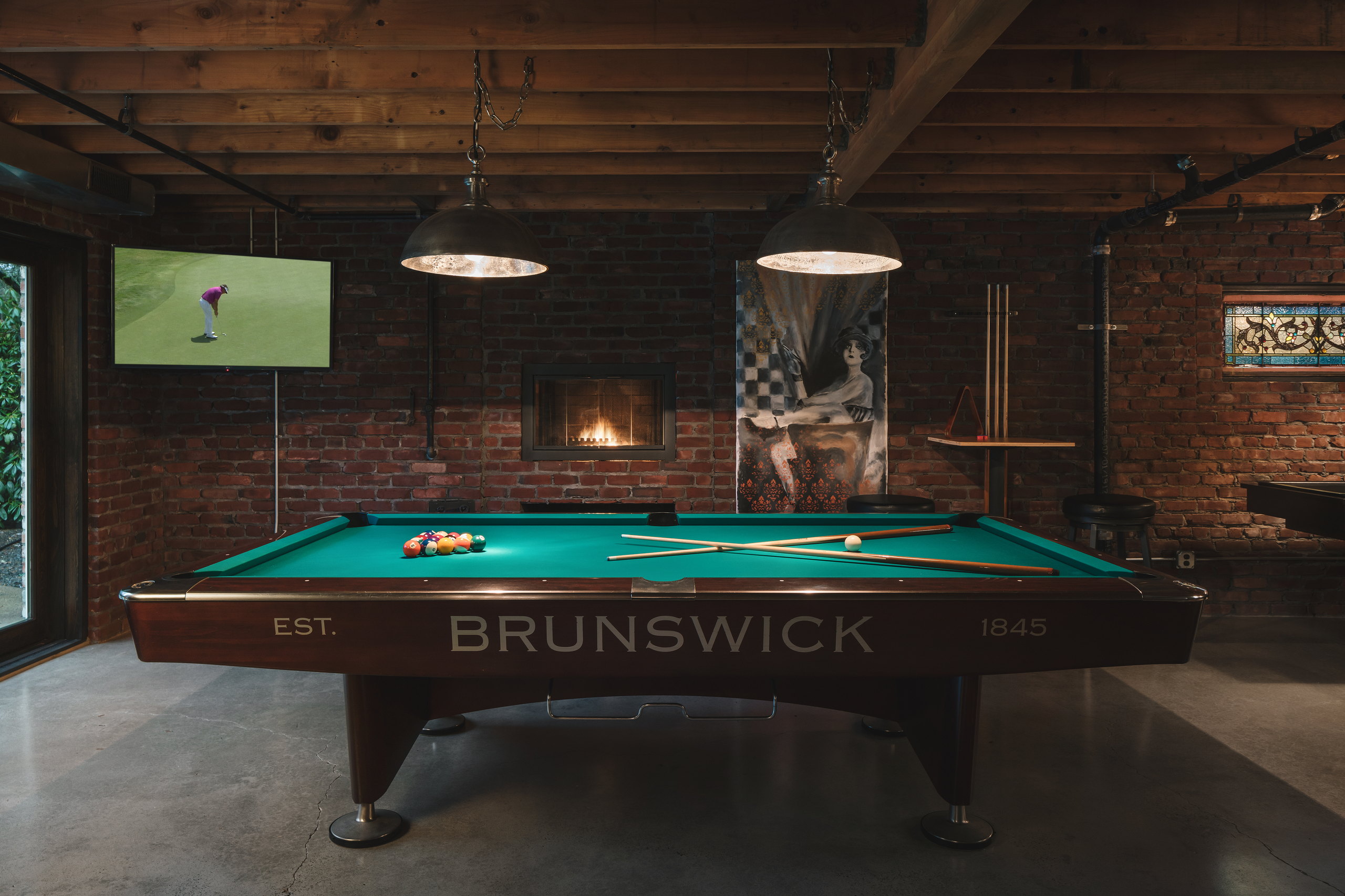 Brunswick pool table lit by two aged-metal pendants in front of an exposed brick wall with a gas fireplace.