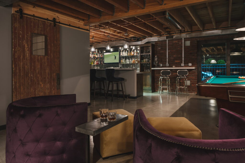 Velvet lounge chairs and leather ottomans facing the bar, with a sliding barn door at left.