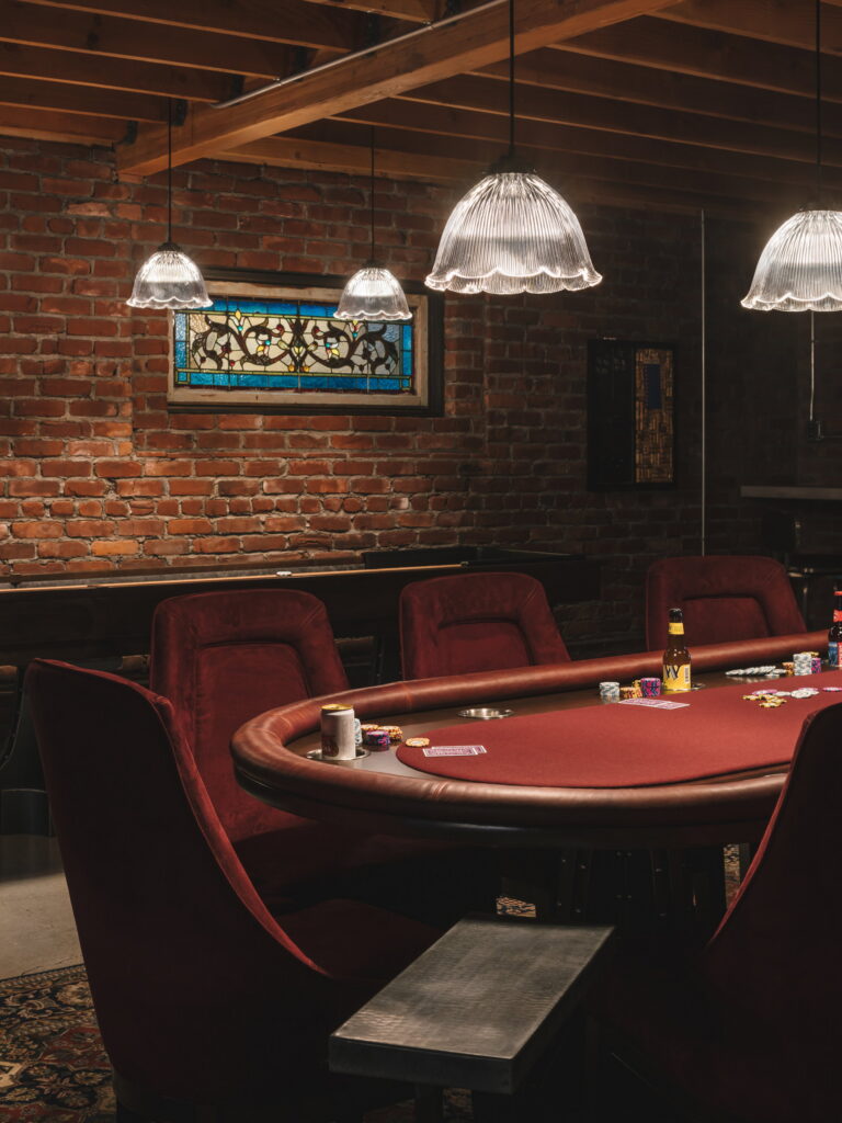 Shuffleboard table on polished concrete beneath two salvaged stained-glass windows set into an exposed brick wall.