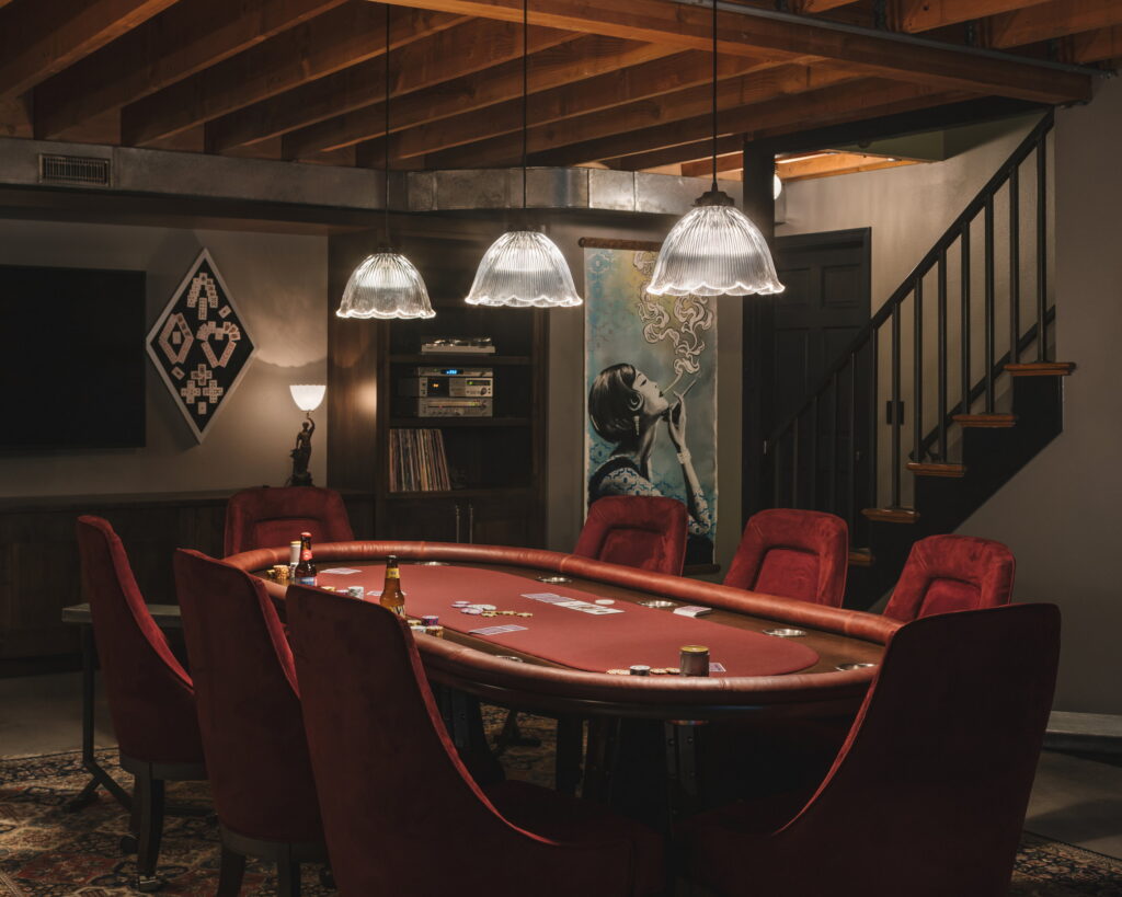 Poker table with red velvet chairs, a record shelf, and the staircase at right.